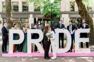 Two grooms and their wedding party standing in front of large letters spelling out PRIDE in Seattle