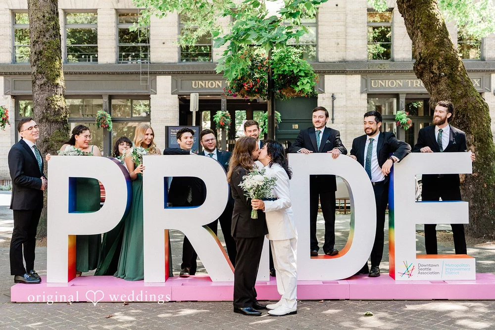 Two grooms and their wedding party standing in front of large letters spelling out PRIDE in Seattle